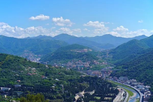 Vista di colline verdi coperte di alberi vicino a Genova, Italia, con antiche fortificazioni e moderne torri di comunicazione sulle cime, sotto un cielo azzurro brillante.