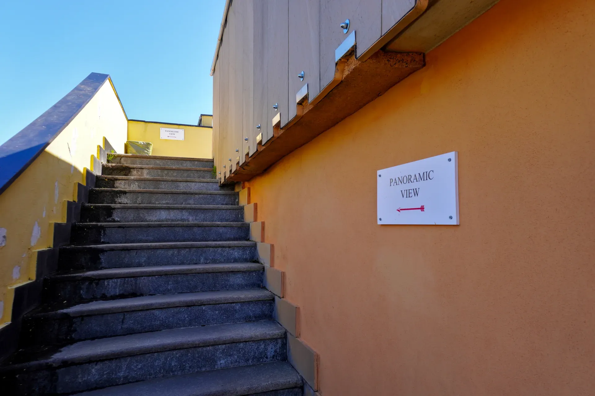Eine Steintreppe führt zu einem Schild mit der Aufschrift 'PANORAMIC VIEW' und einem roten Pfeil nach links, an einer bunten Wand unter einem klaren blauen Himmel in Genua, Italien.