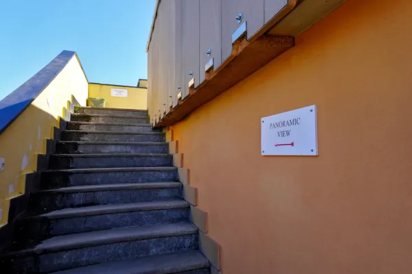 Eine Steintreppe führt zu einem Schild mit der Aufschrift 'PANORAMIC VIEW' und einem roten Pfeil nach links, an einer bunten Wand unter einem klaren blauen Himmel in Genua, Italien.