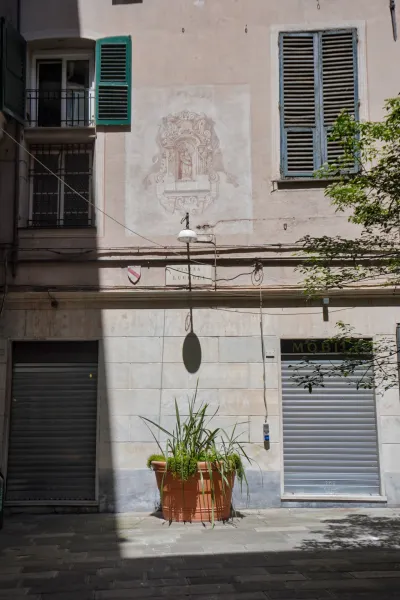 Eine sonnenbeschienene schmale Straße in Genua, Italien, mit Gebäuden mit grünen und blauen Fensterläden, einem verblassten Fresko an der Wand, das eine religiöse Szene darstellt, einer einzelnen Straßenlaterne, einem großen Terrakotta-Topf mit Pflanzen und zwei Rolltoren von Geschäften, die Schatten auf den gepflasterten Boden werfen.