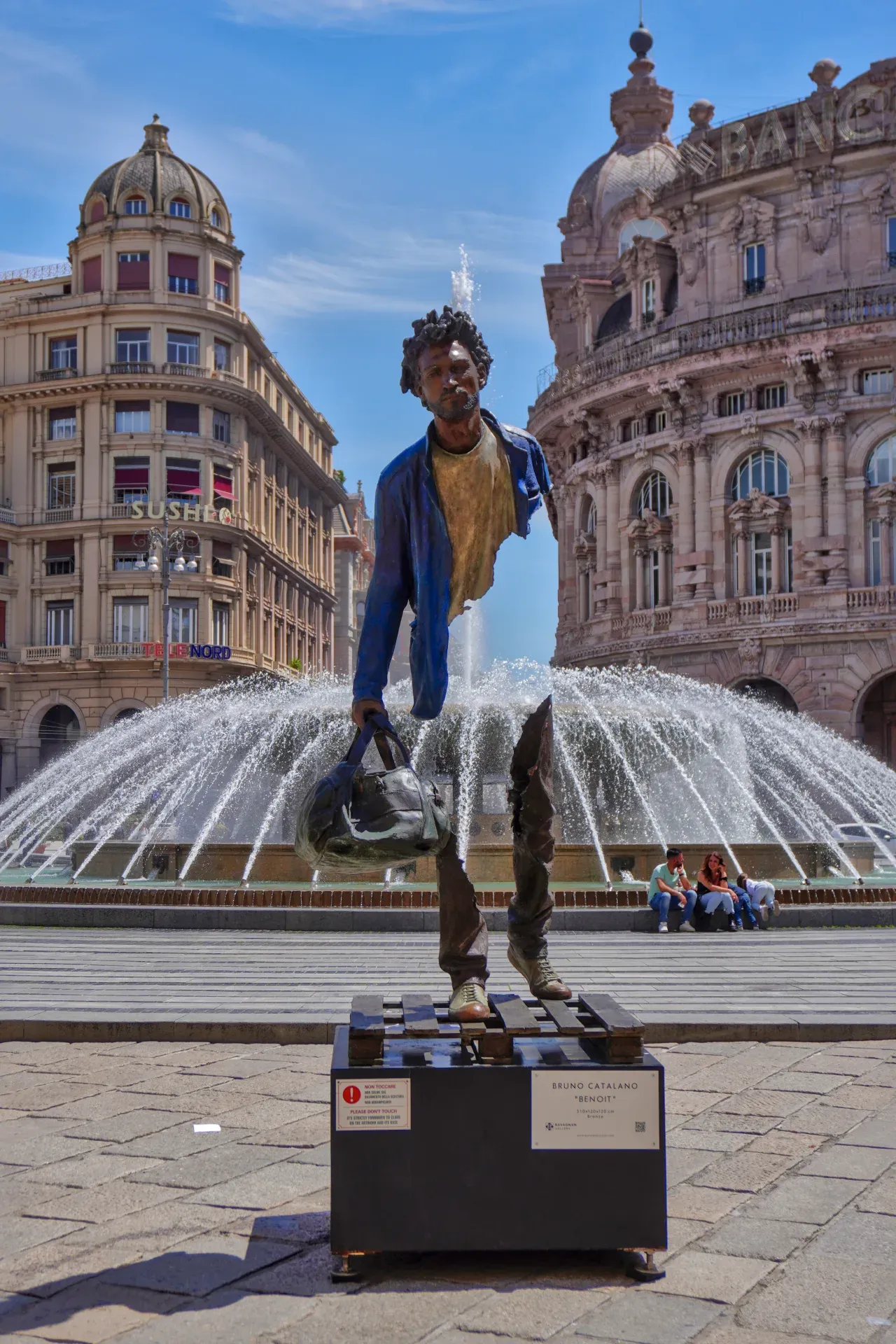 Fragmentovana bronzana skulptura muškarca koji nosi torbu, pod nazivom 'Benoit' Brune Catalana, stoji ispred velikog fontana na gradskom trgu u Đenovi, Italija. Istorijske zgrade su vidljive u pozadini, a ljudi sede pored fontana.