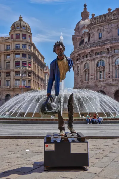 Eine fragmentierte Bronzeskulptur eines Mannes, der eine Tasche trägt, mit dem Titel 'Benoit' von Bruno Catalano, steht vor einem großen Brunnen auf einem Stadtplatz in Genua, Italien. Historische Gebäude sind im Hintergrund sichtbar, und Personen sitzen am Brunnen.