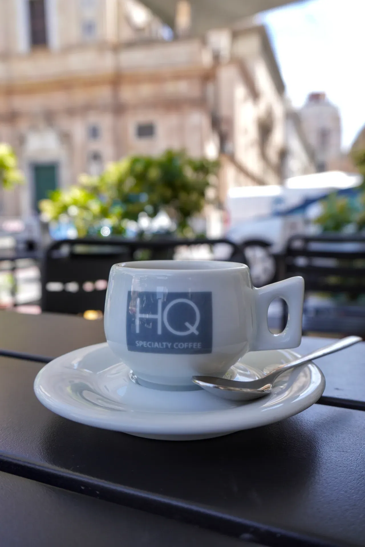 Bela espresso šoljica sa "HQ Specialty Coffee" logom i kašičicom na tacni, na crnom stolu u kafiću na otvorenom u Đenovi, Italija. Pozadina je zamagljena zgradama i zelenilom.