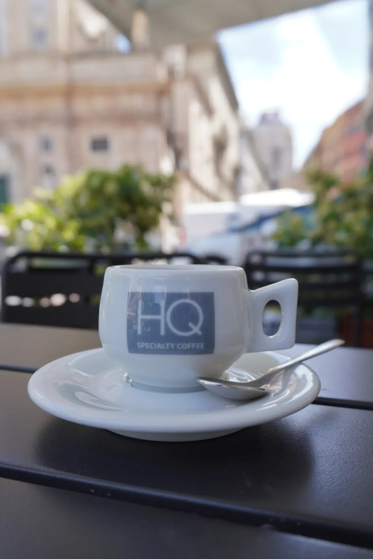Eine weiße Espressotasse mit dem 'HQ Specialty Coffee'-Logo steht auf einer Untertasse mit einem Löffel an einem Außentisch in Genua, Italien, mit unscharfen Gebäuden und Grünflächen im Hintergrund.