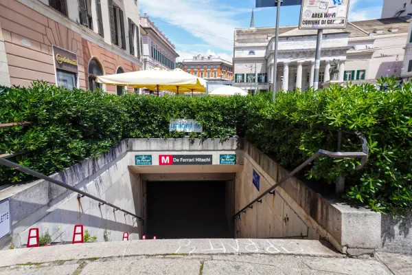 Straßenansicht des Eingangs zur Metrostation De Ferrari in Genua, Italien, mit grünen Hecken, Gebäuden und Beschriftungen.