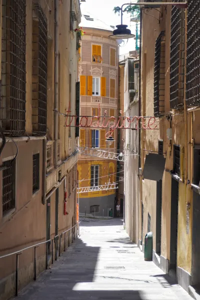 Eine enge Straße in Genua, Italien, mit hohen Gebäuden auf beiden Seiten und Neonschildern mit der Aufschrift 'Tradizione' und 'Curiosità', die über dem Kopfsteinpflaster hängen.