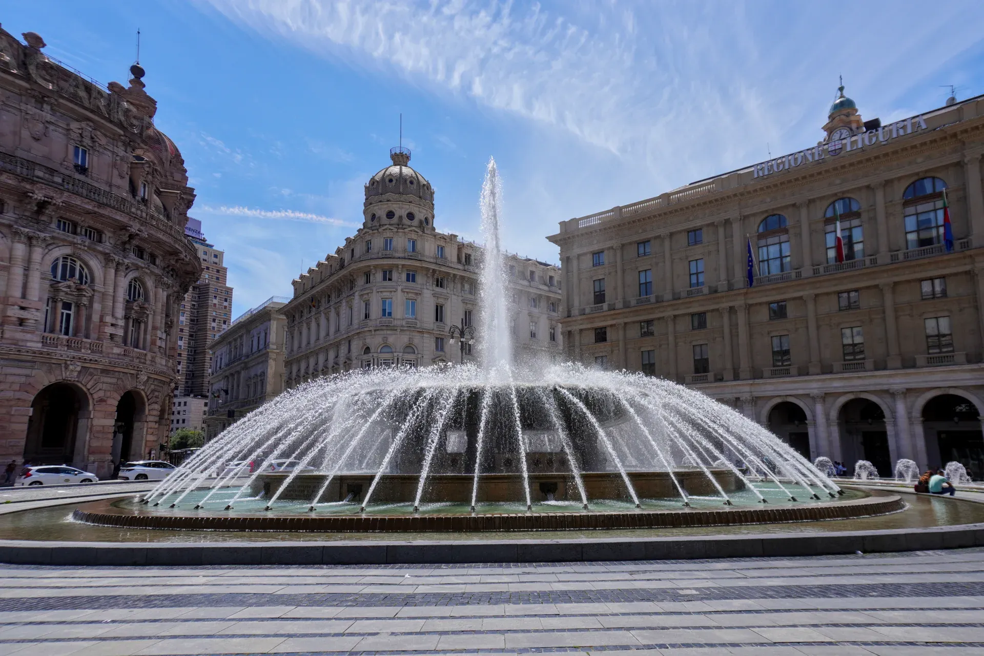 Velika kružna fontana prska vodu u sredini trga, okružena elegantnim istorijskim zgradama u Đenovi, Italija. Nebo iznad je plavo sa dramatičnim perastim oblacima.