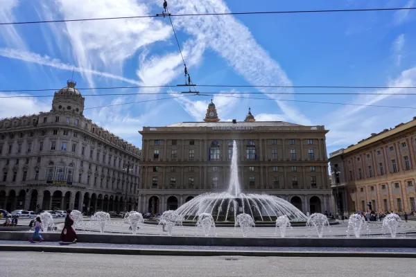 Una grande fontana a più livelli spruzza acqua in Piazza De Ferrari, Genova, Italia. In primo piano, auto bianche, una delle quali è un taxi, e un ciclista sulla strada. Dietro la fontana si trova un magnifico edificio giallo con accenti rosa sotto un cielo blu con nuvole bianche.