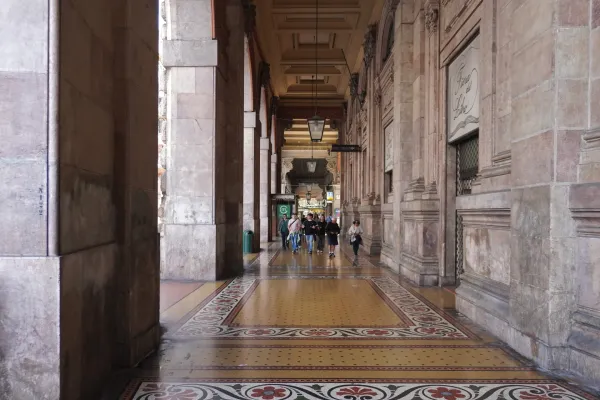 Una prospettiva di un portico coperto a Genova, Italia, con grandi archi in pietra, una scala in marmo e un colorato pavimento in piastrelle di mosaico. La luce del sole proietta ombre sul pavimento, illuminando le decorazioni ornate.