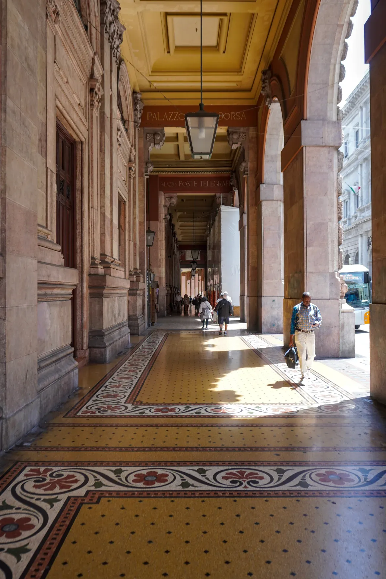 Eine Person geht durch einen breiten, überdachten Gehweg mit hohen Bögen, kunstvollen Säulen und einem gemusterten Fliesenboden in Genua, Italien.