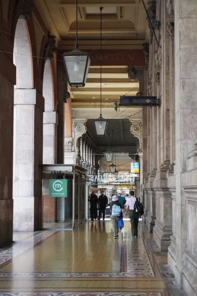 Ein langer, überdachter Gehweg mit gewölbten Säulen führt zum Palazzo della Borsa. Der Boden ist mit bunten Mosaikfliesen verziert, und Sonnenlicht fällt herein und wirft Schatten. Mehrere Personen gehen vom Betrachter weg den Portikus entlang.