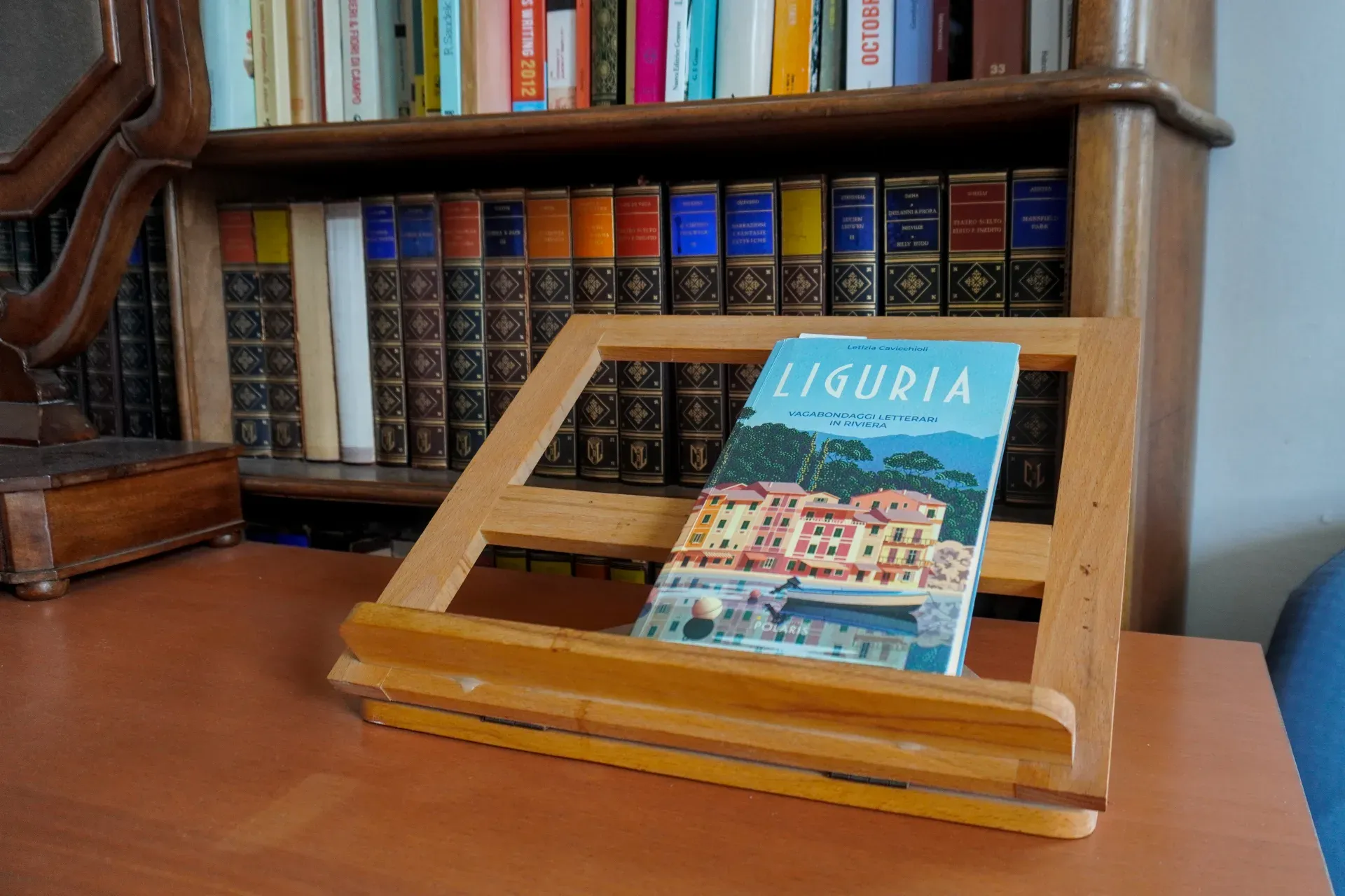 Ein Buch mit dem Titel 'Ligurien' wird auf einer Holzstaffelei präsentiert, die auf einem Schreibtisch vor einem großen Bücherregal mit antiken Büchern steht. Der Buchumschlag zeigt einen malerischen Blick auf die ligurische Architektur und ein Boot.