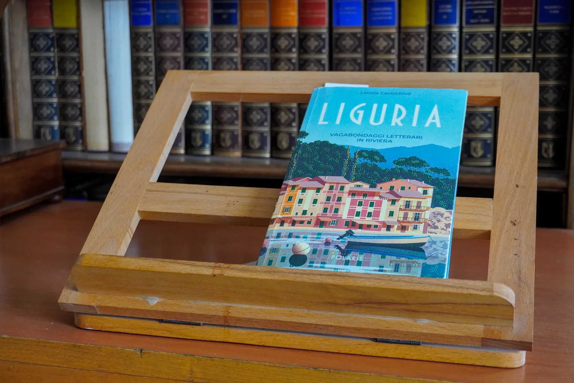 Ein Buch mit einer bunten Illustration von ligurischen Küstenhäusern und einem Boot auf einem Holzständer, mit einem Regal mit alten Büchern im Hintergrund.