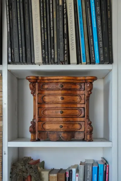 Eine Nahaufnahme einer kleinen, antiken Holzkommode mit fünf Schubladen, jede mit einem kleinen runden Knopf, auf einem weißen Regal. Über der Kommode ist eine Reihe von CD-Hüllen mit Titeln klassischer Musik sichtbar. Unter der Kommode enthält ein weiteres Regal eine Vielzahl von Büchern und ein dekoratives Metallstück.