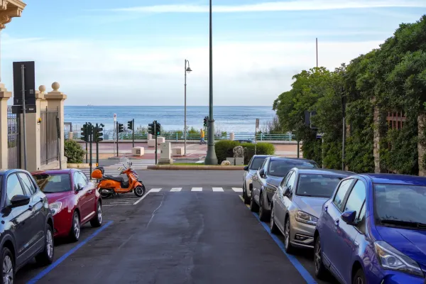 Eine Straße in Genua, Italien, mit Autos, die an den Rändern geparkt sind, und dem Meer im Hintergrund, eingerahmt von Gebäuden und Bäumen.