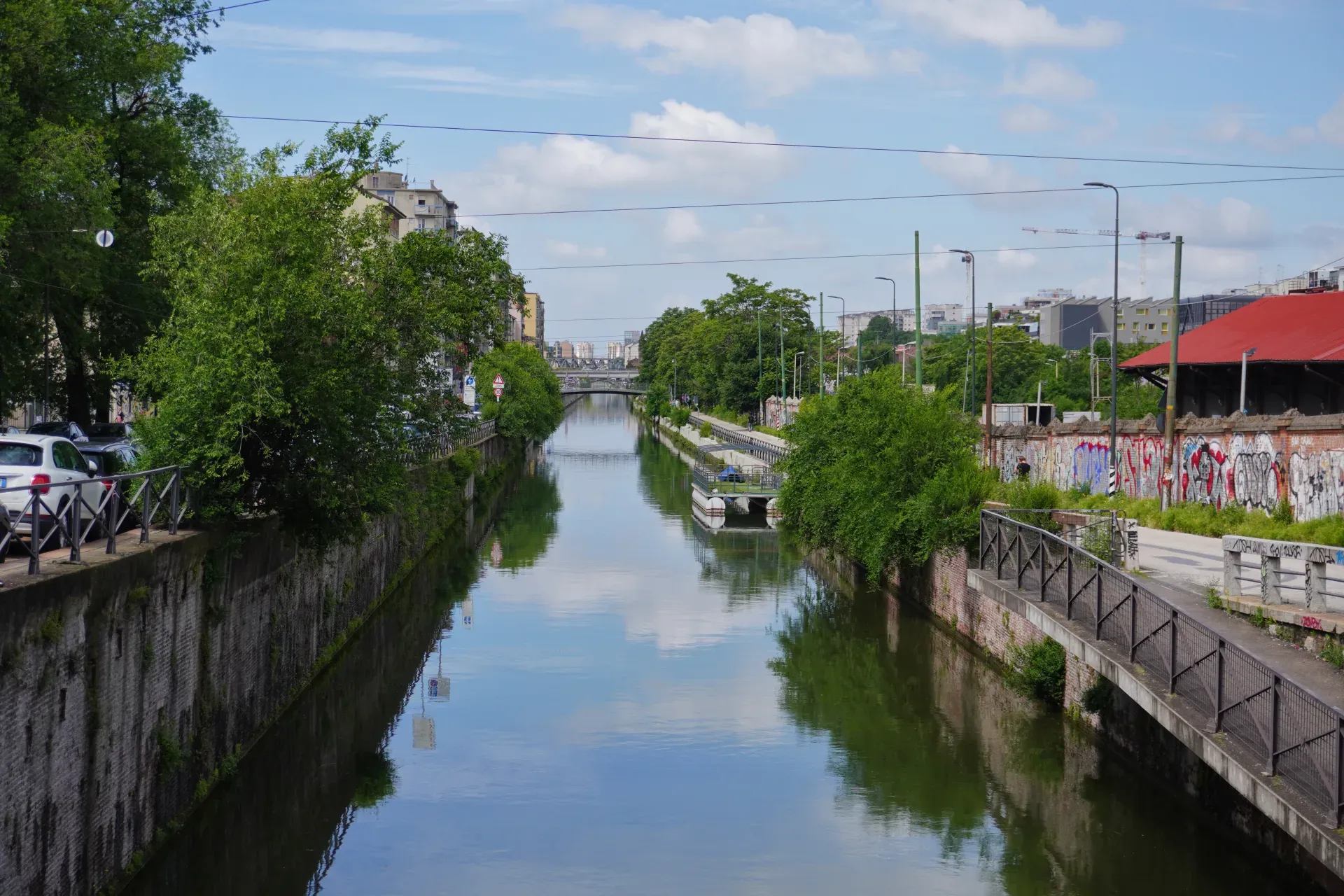 Širok kanal u Milanu, Italija, sa zelenim drvećem koje obrubljuje obale, kamenim zidovima i modernim zgradama u pozadini. Nebo i drveće se odražavaju u mirnoj vodi, a most premošćuje kanal u daljini.