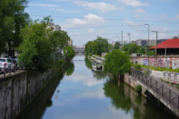 Un ampio canale a Milano, Italia, con alberi verdi che costeggiano le sponde, mura di pietra ed edifici moderni sullo sfondo. Il cielo e gli alberi si riflettono nell'acqua calma, e un ponte attraversa il canale in lontananza.