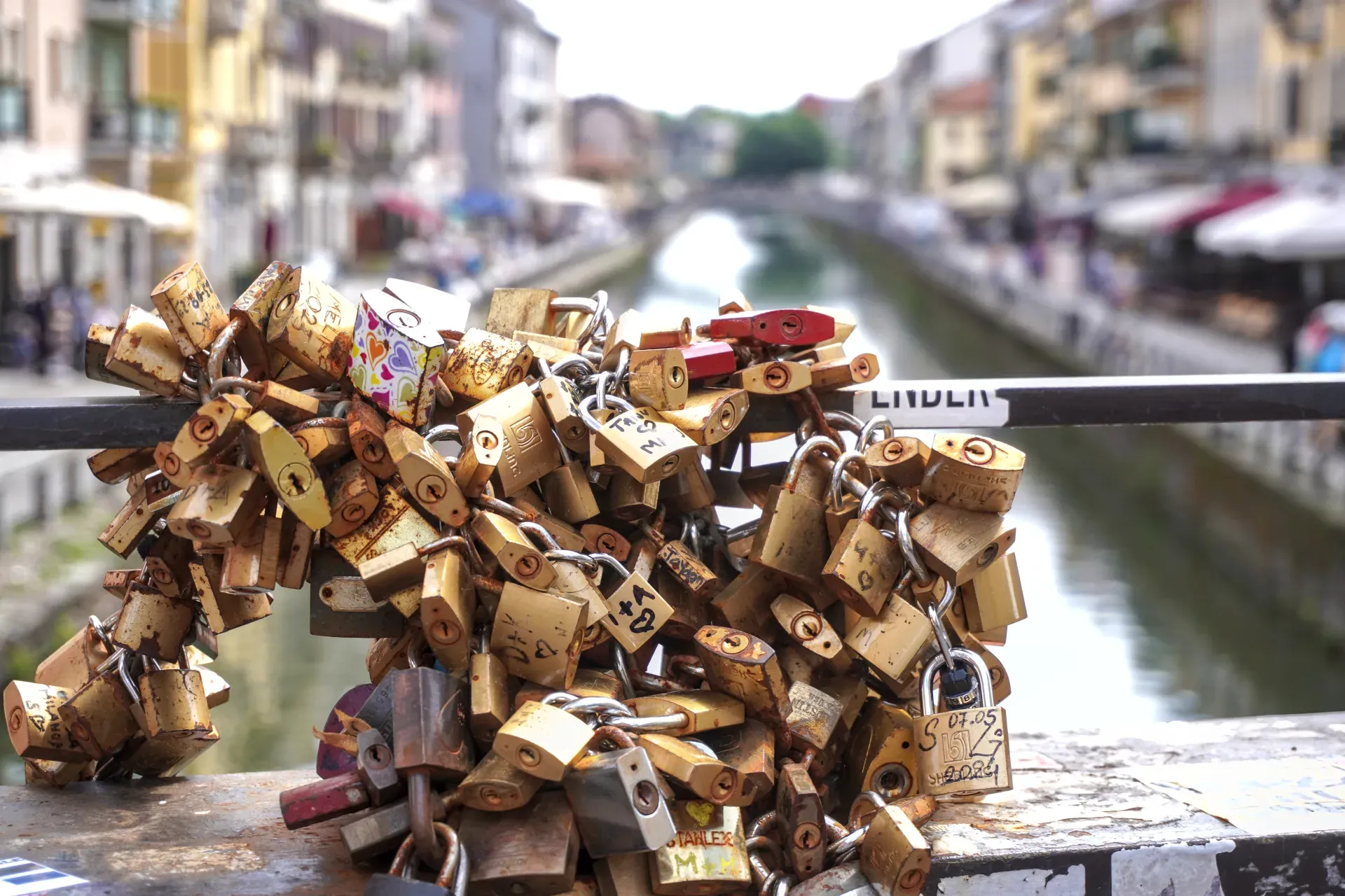 Una pila di lucchetti usurati e colorati attaccati a catene sul parapetto di un ponte, con un canale e gli edifici di Milano sfocati sullo sfondo.
