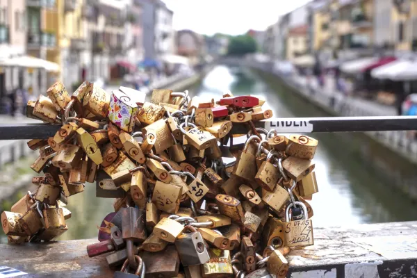Una pila di lucchetti usurati e colorati attaccati a catene sul parapetto di un ponte, con un canale e gli edifici di Milano sfocati sullo sfondo.