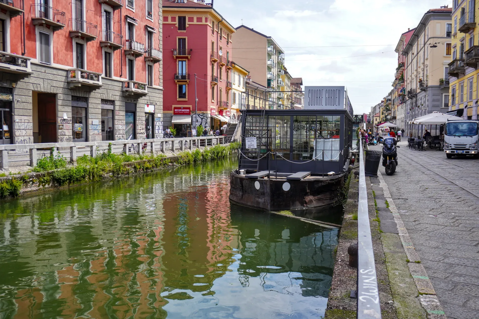 Kanal u milanskom kvartu Navigli sa šarenim zgradama sa obe strane, pešačkom stazom, ljudima koji šetaju, čamcem na vodi i mostom u pozadini.