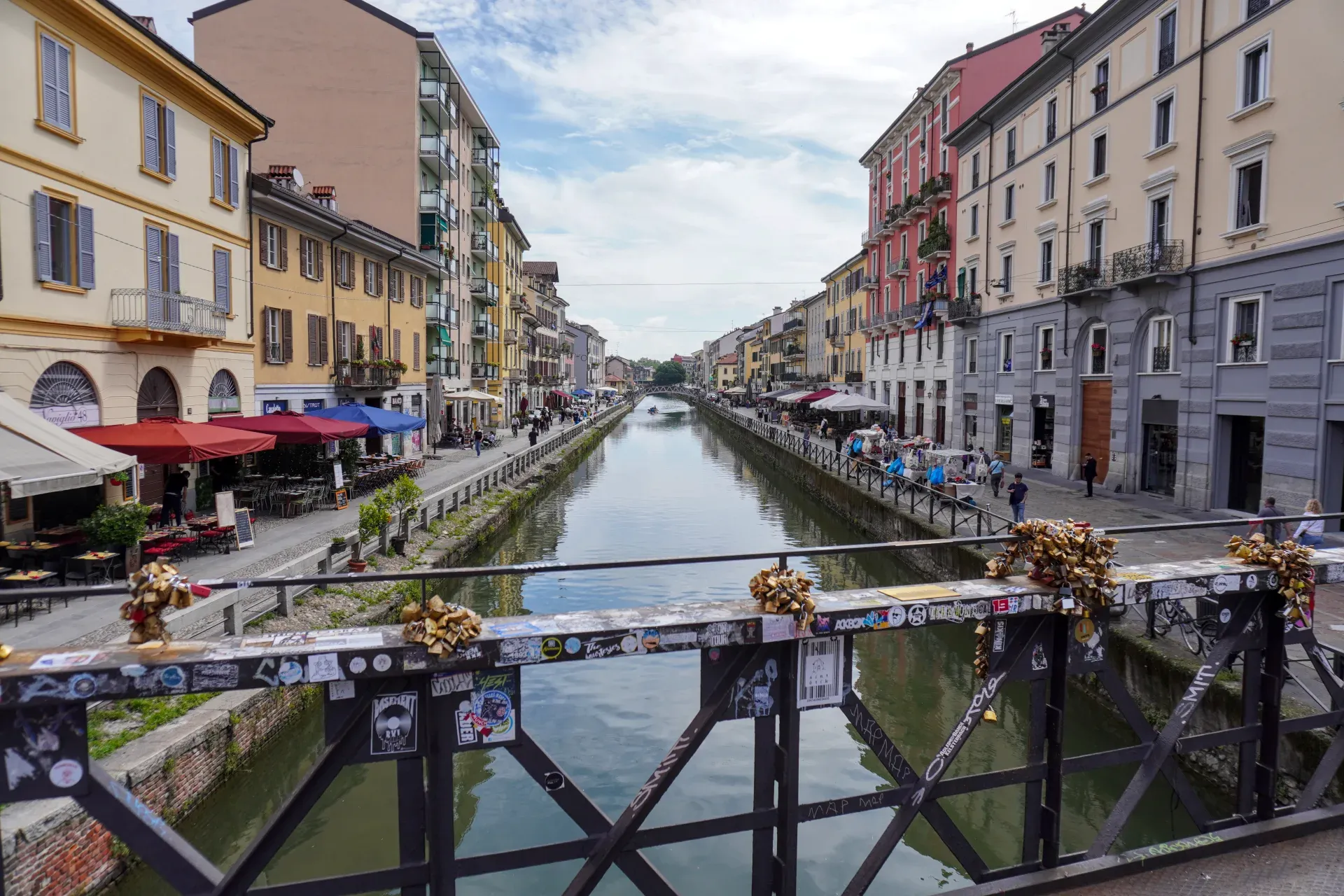 Menschen rudern mit Booten auf einem Kanal, der von historischen Gebäuden und Außenrestaurants im Navigli-Viertel Mailands gesäumt ist.