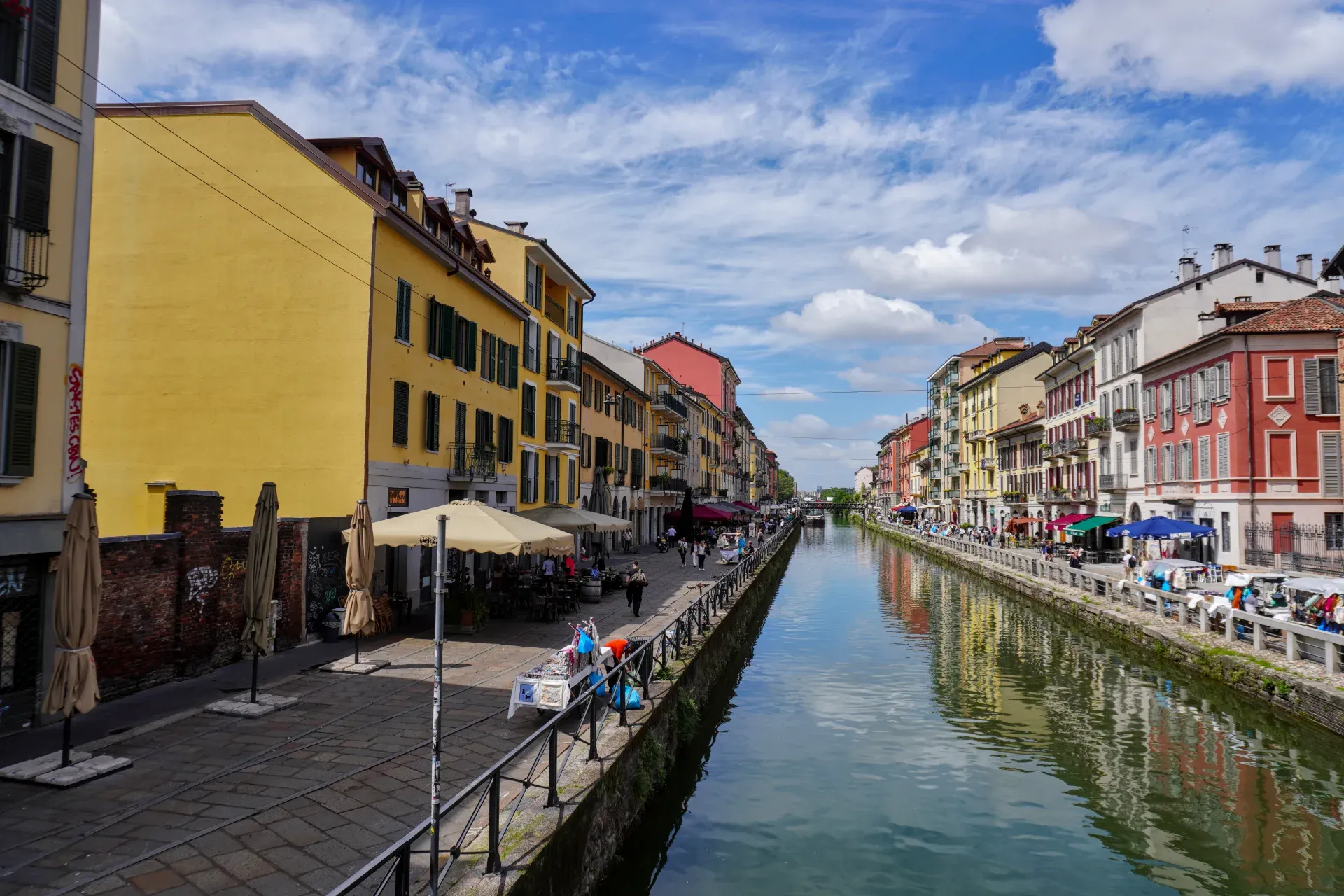Una giornata di sole nel quartiere Navigli di Milano, che mostra un canale con edifici e persone su barche.