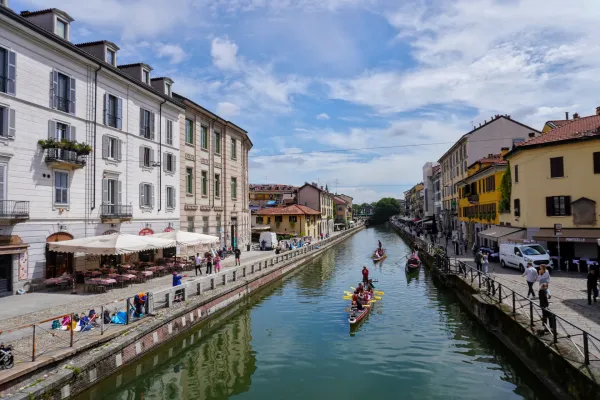 Un ampio canale nel quartiere Navigli di Milano, fiancheggiato da edifici gialli e beige, con un ponte pedonale che lo attraversa. Barche sono ormeggiate nel canale e persone sono visibili sul ponte e lungo il camminamento del canale.