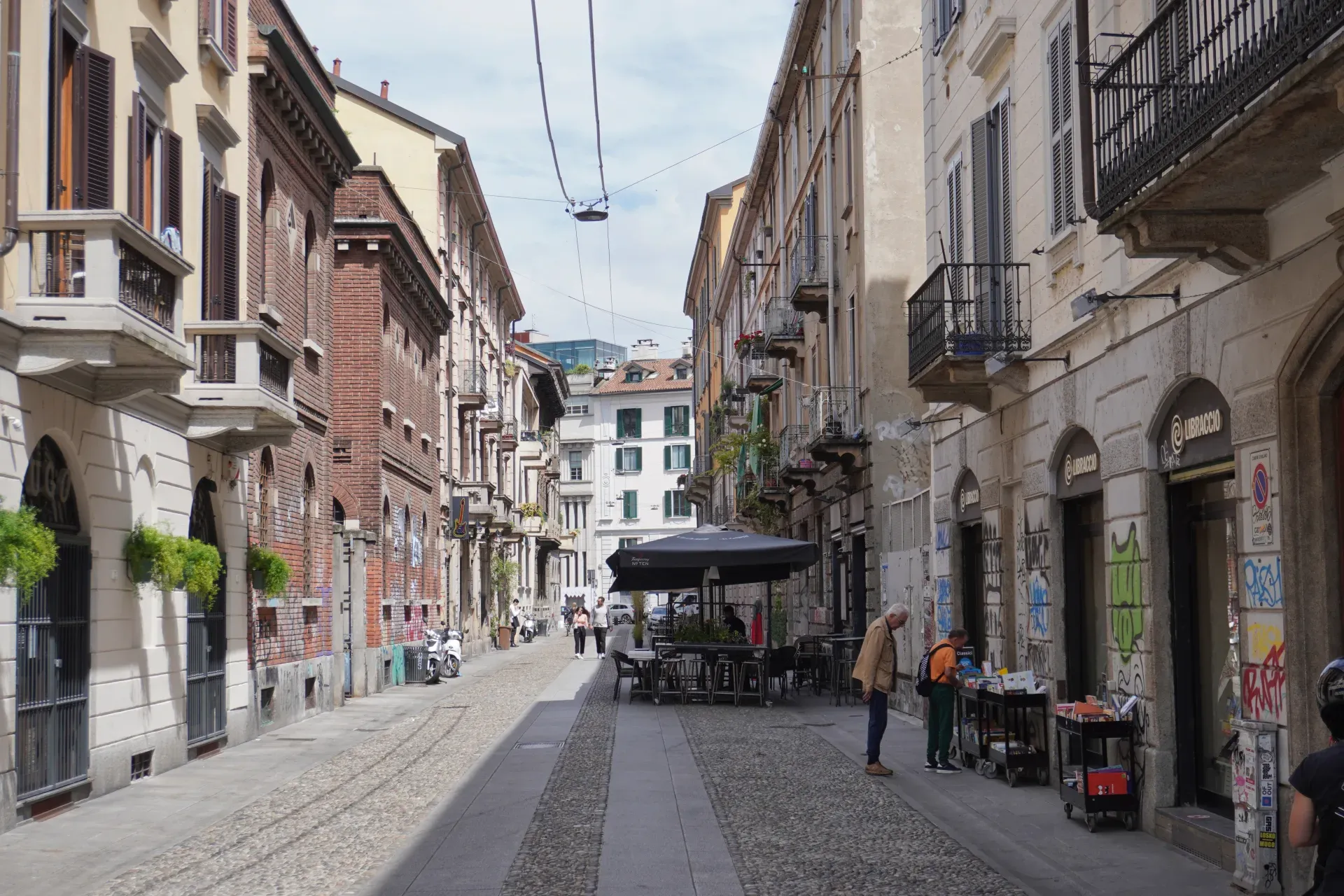 Ein Mann auf einem Fahrrad überquert eine Fußgängerstraße in Mailand, Italien. Gebäude mit Fensterläden, Balkonen, Graffiti und Geschäften säumen die Gasse.