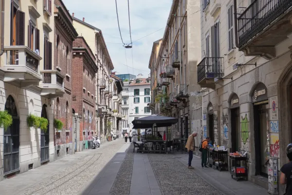 Un uomo in bicicletta attraversa una strada pedonale a Milano, Italia. Edifici con persiane, balconi, graffiti e negozi fiancheggiano il vicolo.
