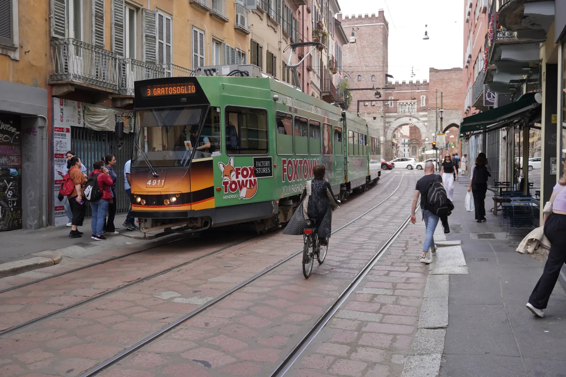 Eine gelbe Straßenbahn auf einer Kopfsteinpflasterstraße in Mailand mit Gebäuden und Cafés an den Seiten.