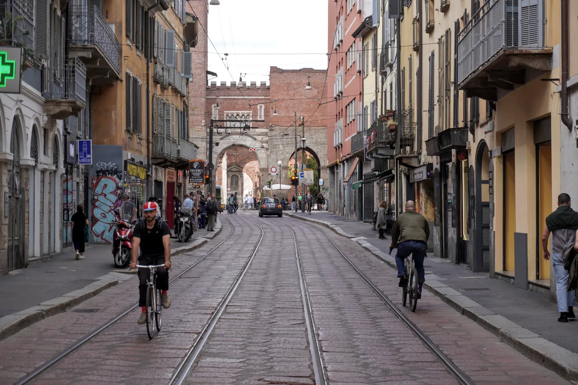 Un tram verde con marchio 'Foxtown' percorre una strada di Milano, Italia, con pedoni sul marciapiede e un ciclista nelle vicinanze. Edifici storici e un arco sono visibili sullo sfondo.