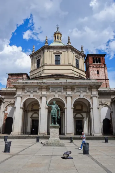 Statua dell'imperatore Costantino davanti alla Basilica di San Lorenzo Maggiore con cupola e campanile sotto un cielo nuvoloso.