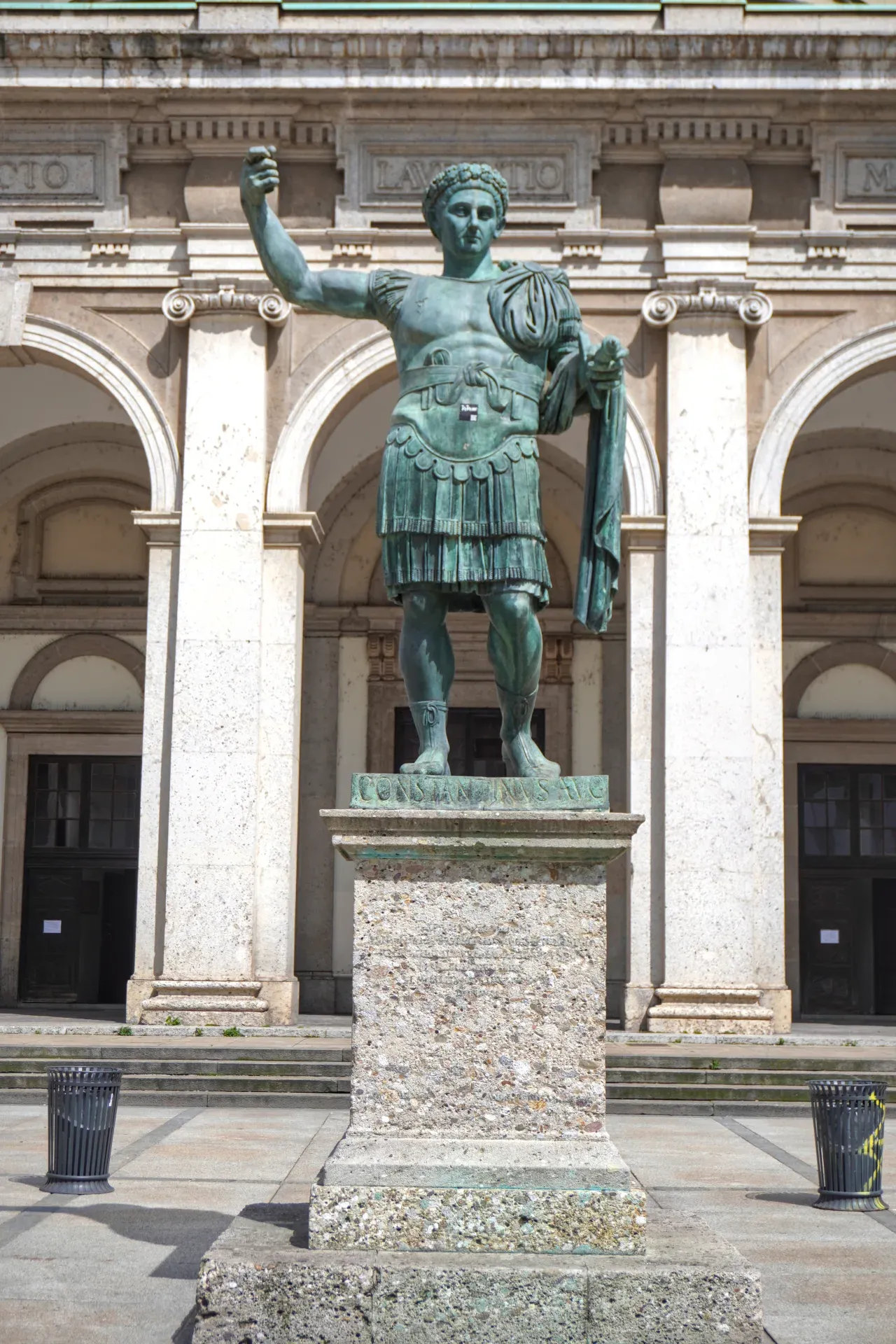 Un'alta statua in bronzo dell'imperatore Costantino il Grande in armatura militare, in piedi su un piedistallo di pietra di fronte a un edificio con grandi archi e architettura classica a Milano, Italia.