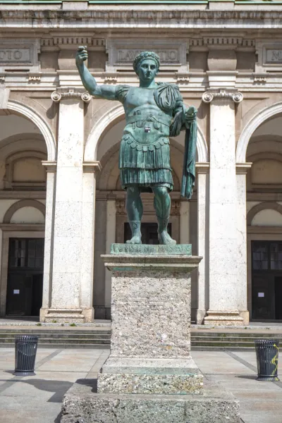 Eine hohe Bronzestatue von Kaiser Konstantin dem Großen in militärischer Rüstung, stehend auf einem Steinsockel vor einem Gebäude mit großen Bögen und klassischer Architektur in Mailand, Italien.