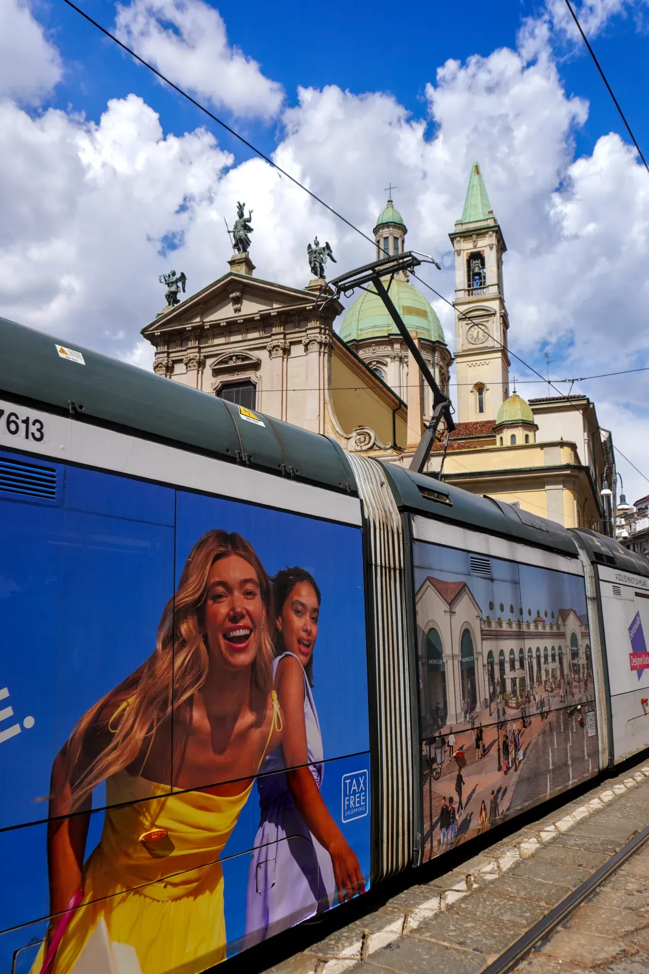 Eine Straßenbahn in Mailand, Italien, mit bunten Werbetafeln an den Seiten, fährt an einer Kirche und historischen Gebäuden unter einem hellen, bewölkten Himmel vorbei.