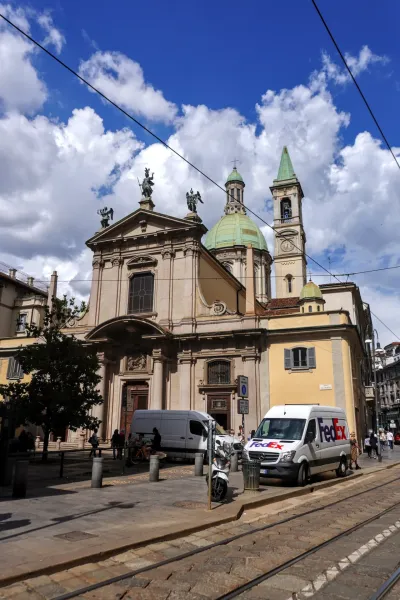 Eine europäische Kirche mit Kuppel und klassischer Architektur steht an einer Straße in Mailand, mit Straßenbahnschienen im Vordergrund und einem geparkten FedEx-Lieferwagen unter einem blauen Himmel mit Wolken.