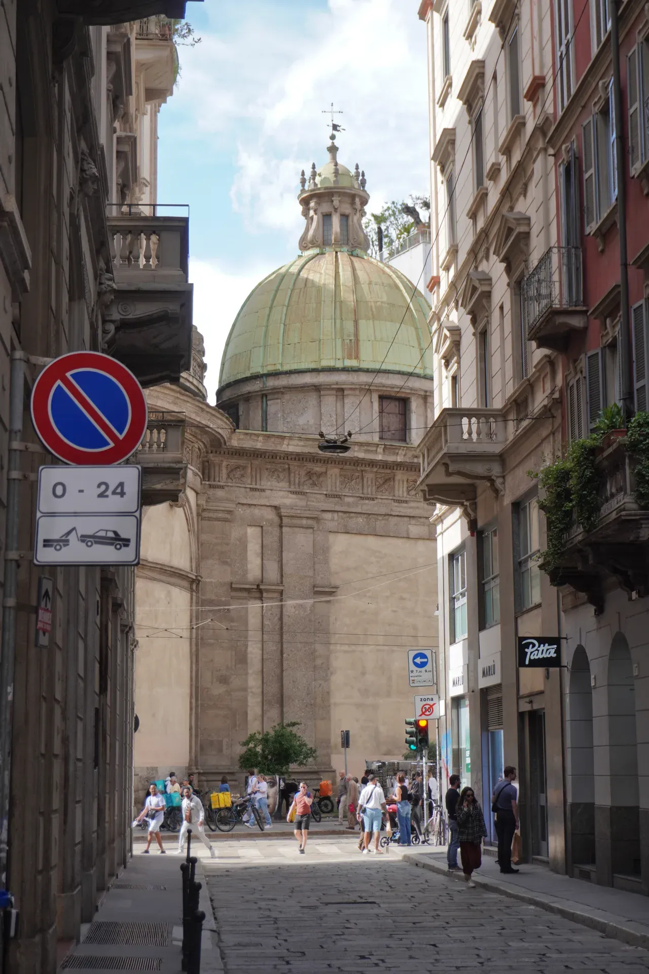 Eine europäische Straße in Mailand, Italien, mit einer großen grünen Kuppel, die zwischen Gebäuden sichtbar ist, und Menschen, die auf einer Kopfsteinpflasterstraße gehen.
