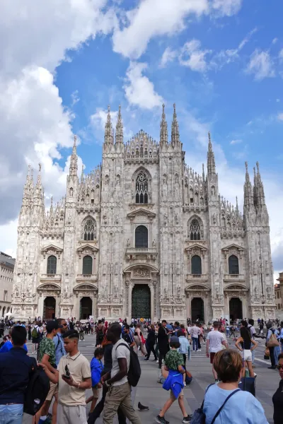 La facciata ornata del Duomo di Milano, Italia, con molte guglie e dettagli in pietra sotto un cielo blu brillante con nuvole sparse. Una folla di persone è visibile in primo piano sulla piazza.