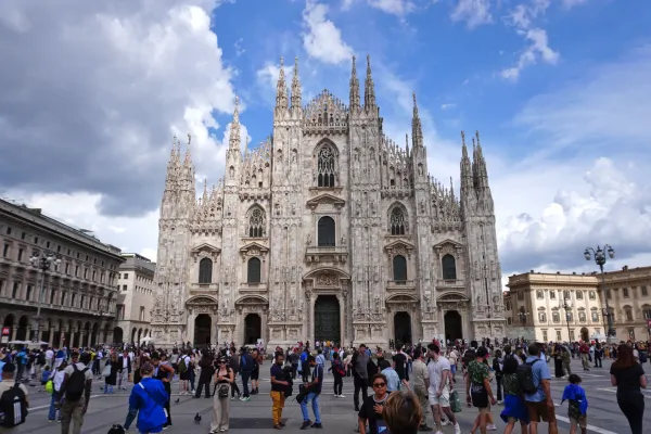 Una veduta d'insieme del Duomo di Milano, una grande cattedrale gotica con numerose guglie e intricata lavorazione in pietra, su uno sfondo di cielo nuvoloso. Persone sono visibili sulla piazza in primo piano.