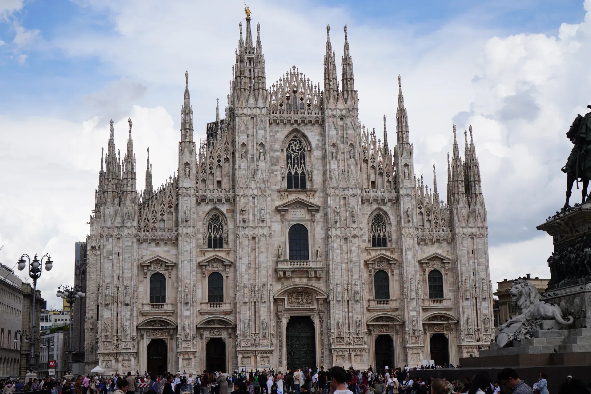 Die aufwendige Fassade des Mailänder Doms (Duomo di Milano) unter einem bewölkten Himmel, mit einer bronzenen Reiterstatue und Bäumen davor sowie einer Menschenmenge auf dem Platz.