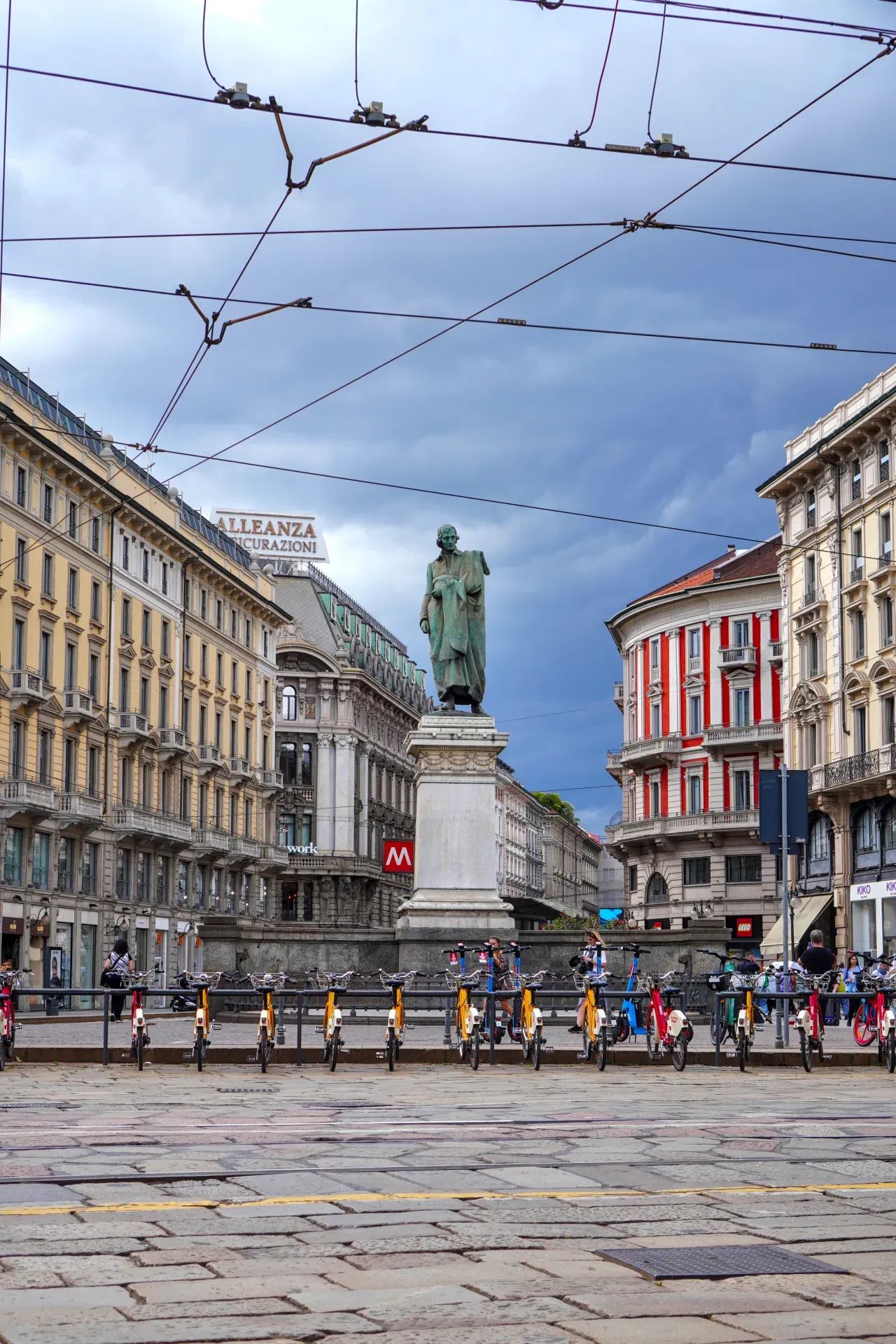 Eine Totale der Piazza Cavour in Mailand, Italien, dominiert von einer Bronzestatue auf einem Sockel, mit verzierten Gebäuden, die den Platz säumen, und einer Reihe von Leihfahrrädern im Vordergrund.