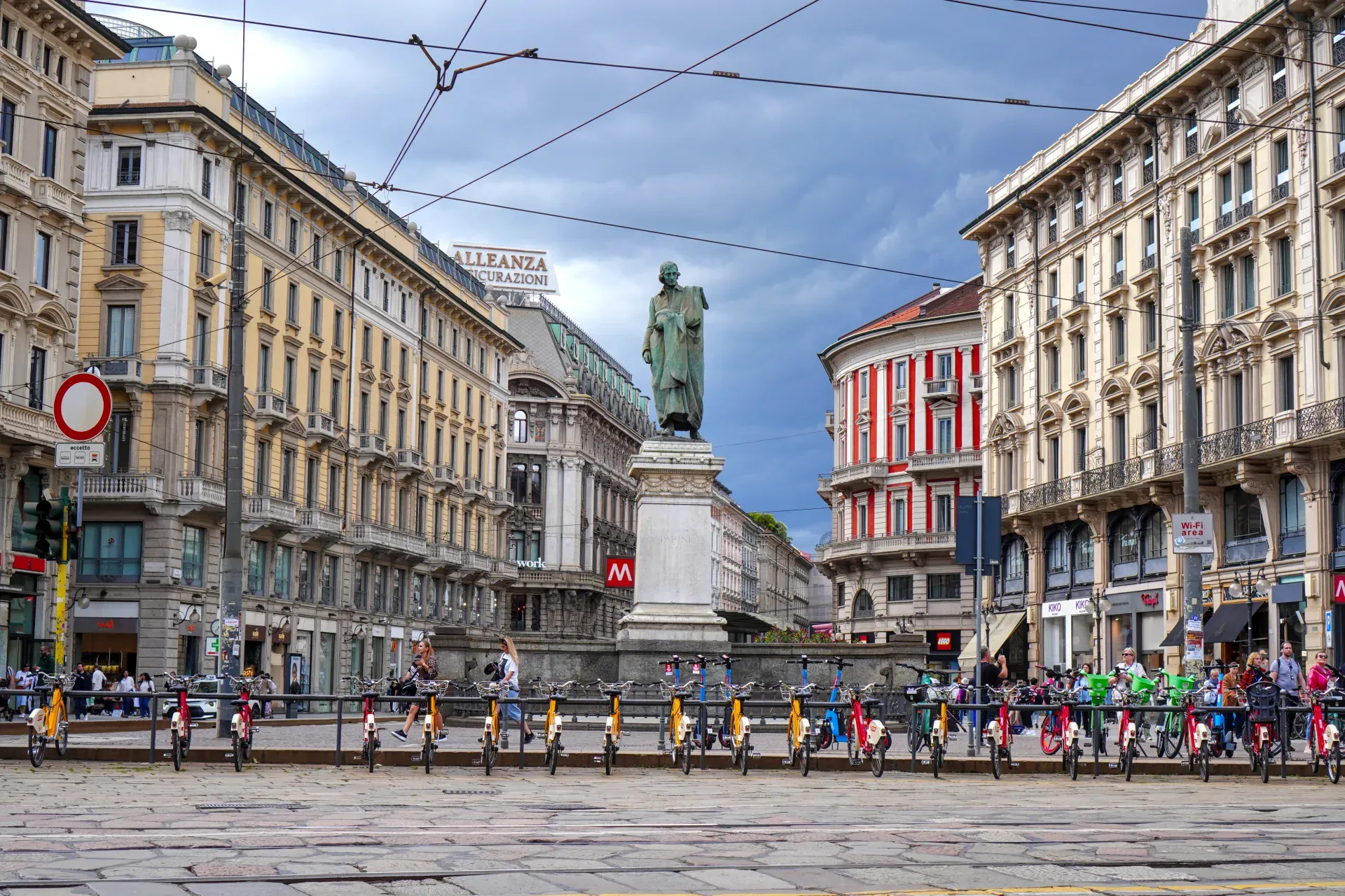 Ulica u Milanu, Italija, sa ukrašenim zgradama koje je okružuju, bronzanom statuom na postolju u sredini i brojnim žutim i crvenim električnim trotinetima i biciklama parkiranim u nizu.