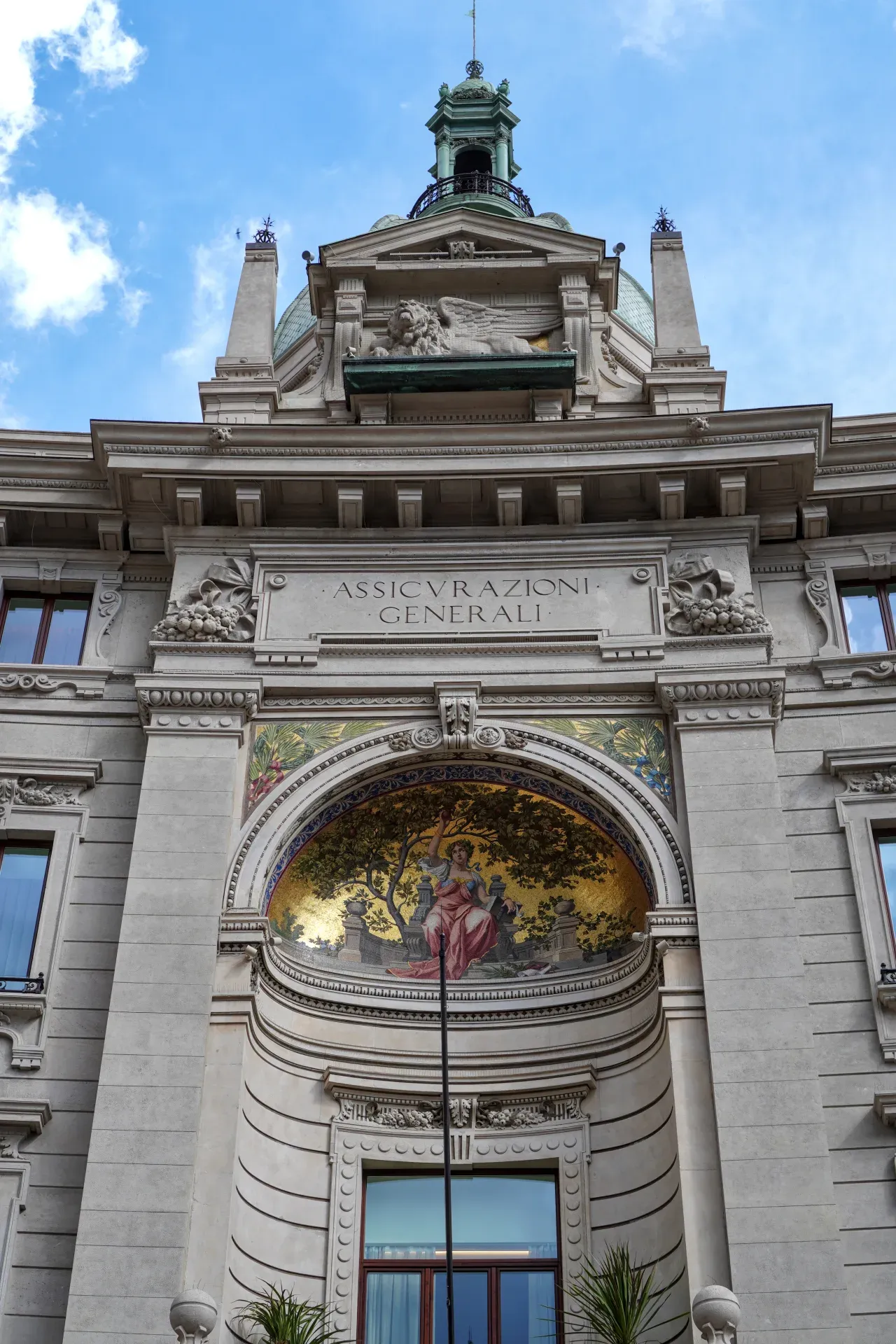 Una vista dettagliata della facciata superiore dell'edificio Assicurazioni Generali a Milano, che mette in mostra elementi architettonici classici, un mosaico e un leone alato.