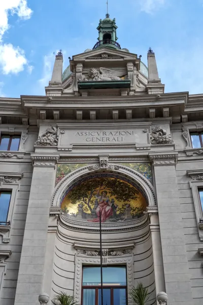 Una vista dettagliata della facciata superiore dell'edificio Assicurazioni Generali a Milano, che mette in mostra elementi architettonici classici, un mosaico e un leone alato.
