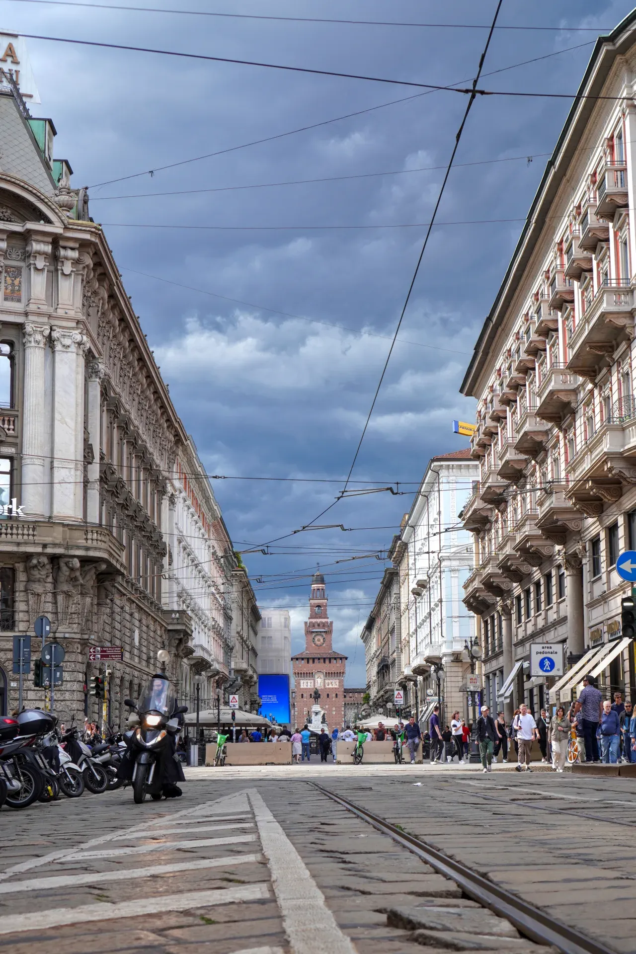 Una strada a Milano, Italia, con un tram che corre su binari di ciottoli, fiancheggiata da edifici storici. Il Castello Sforzesco è visibile sullo sfondo sotto un cielo nuvoloso.