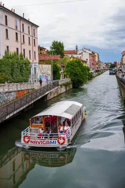 Un battello turistico chiamato "Navigami" naviga su un canale a Milano, Italia, con passeggeri a bordo. Edifici e spazi verdi costeggiano il canale e il riflesso della barca è visibile nell'acqua.