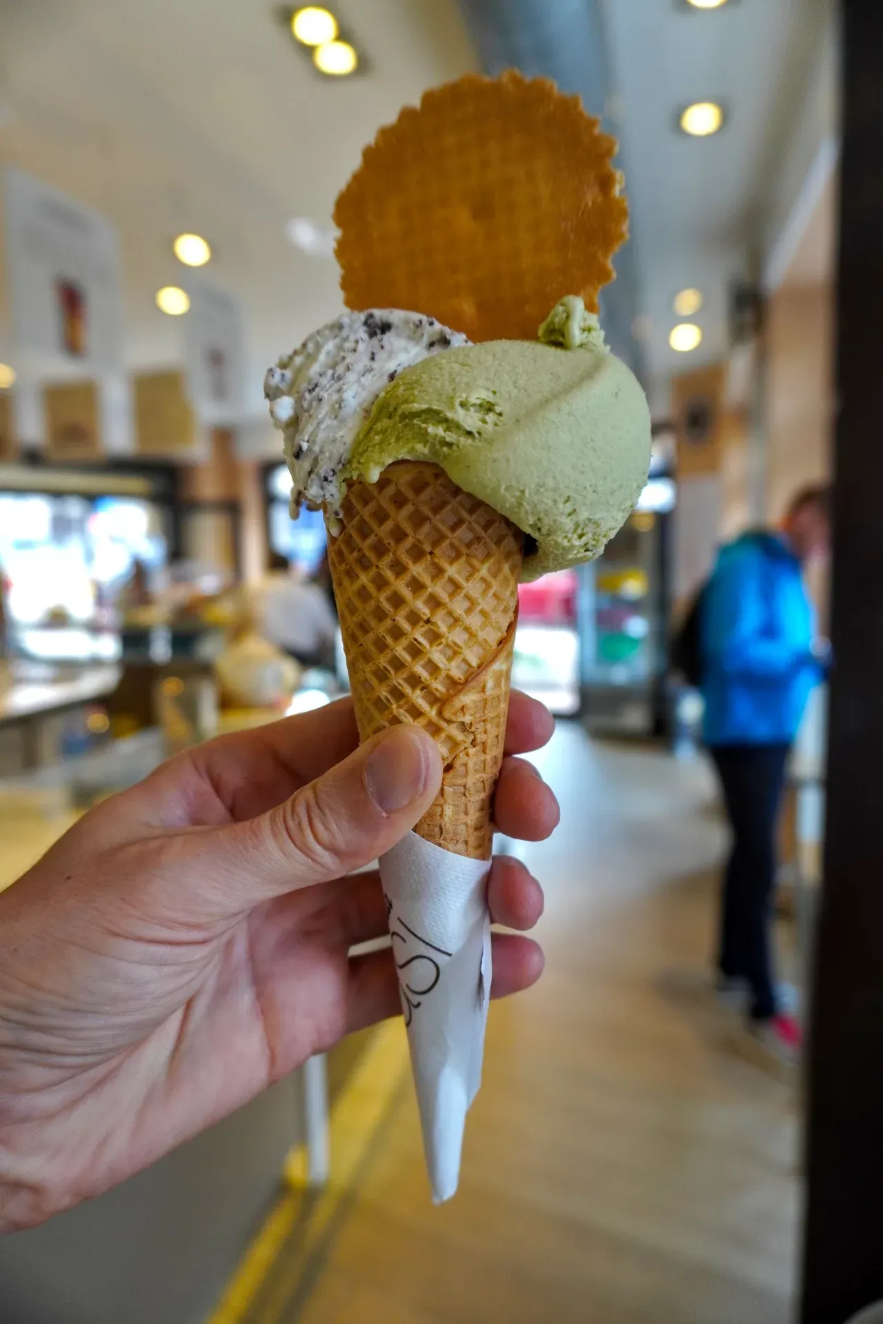 Una mano tiene un cono gelato con tre palline di gelato, tra cui pistacchio e crema biscotto, guarnito con una cialda rotonda. Nello sfondo sfocato si vedono persone all'interno di una gelateria a Milano.