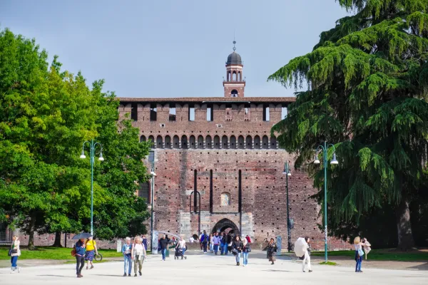 Menschen spazieren vor dem Schloss Sforzesco in Mailand, Italien, mit großen Bäumen, die die Aussicht umrahmen.