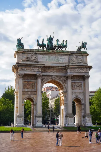 L'imponente Arco della Pace a Milano, Italia, sotto un cielo nuvoloso, con persone in primo piano e alberi che circondano il monumento.