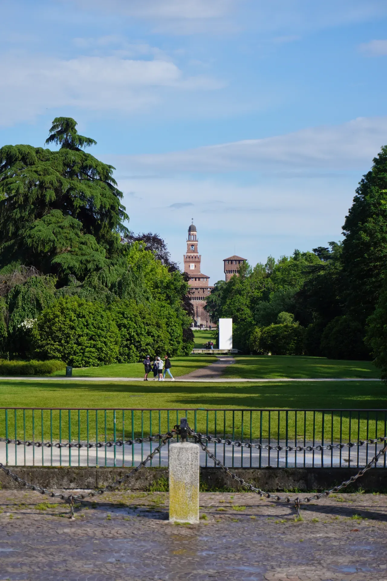 La torre del Castillo Sforza se eleva sobre los árboles en el Parco Sempione, Milán, con personas caminando por un sendero en primer plano.